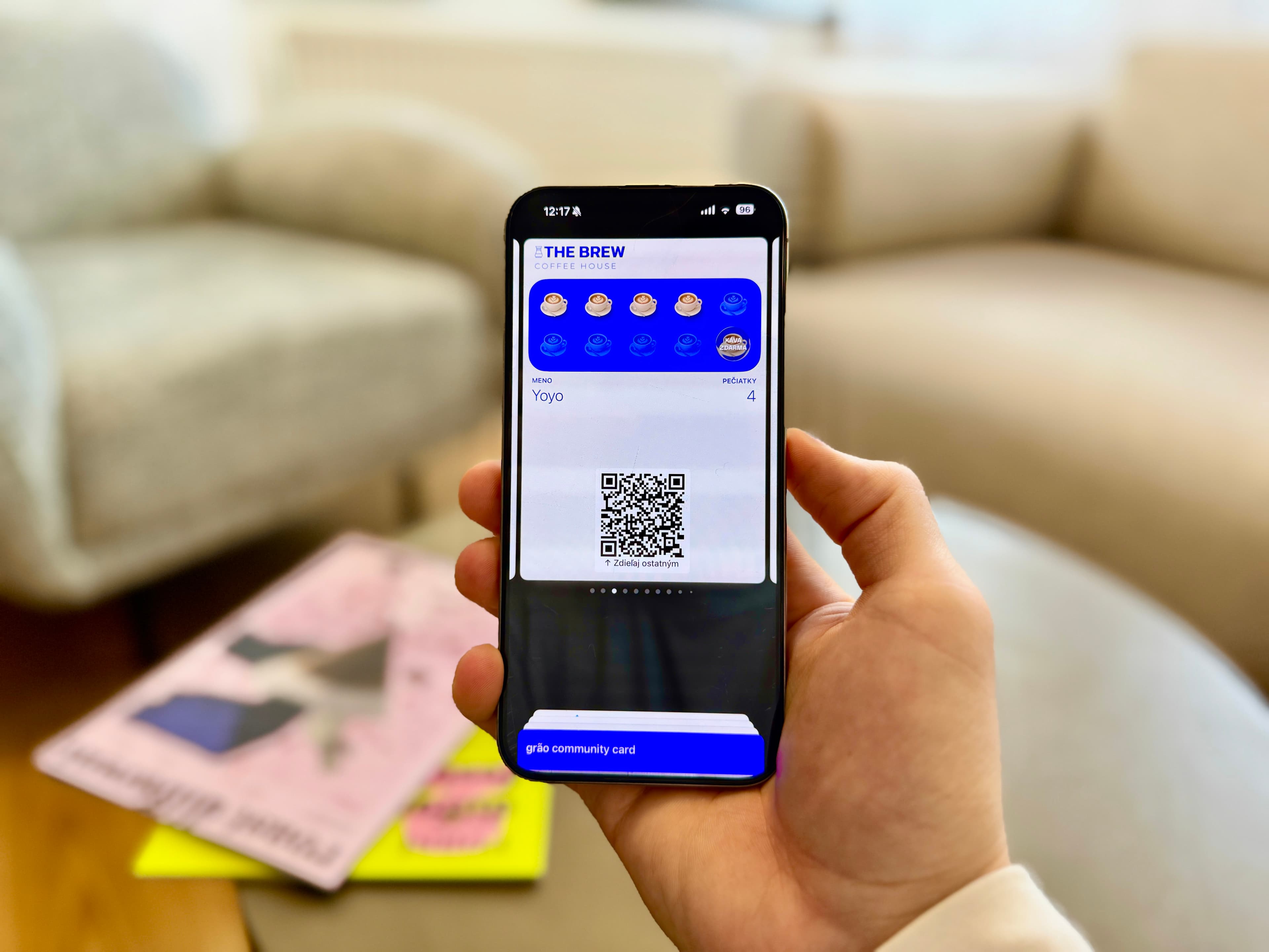 Ruka drží smartfón zobrazujúci digitálnu vernostnú kartu kaviarne s ikonami pečiatok a QR kódom, pričom v pozadí je rozmazaný stôl s časopismi.