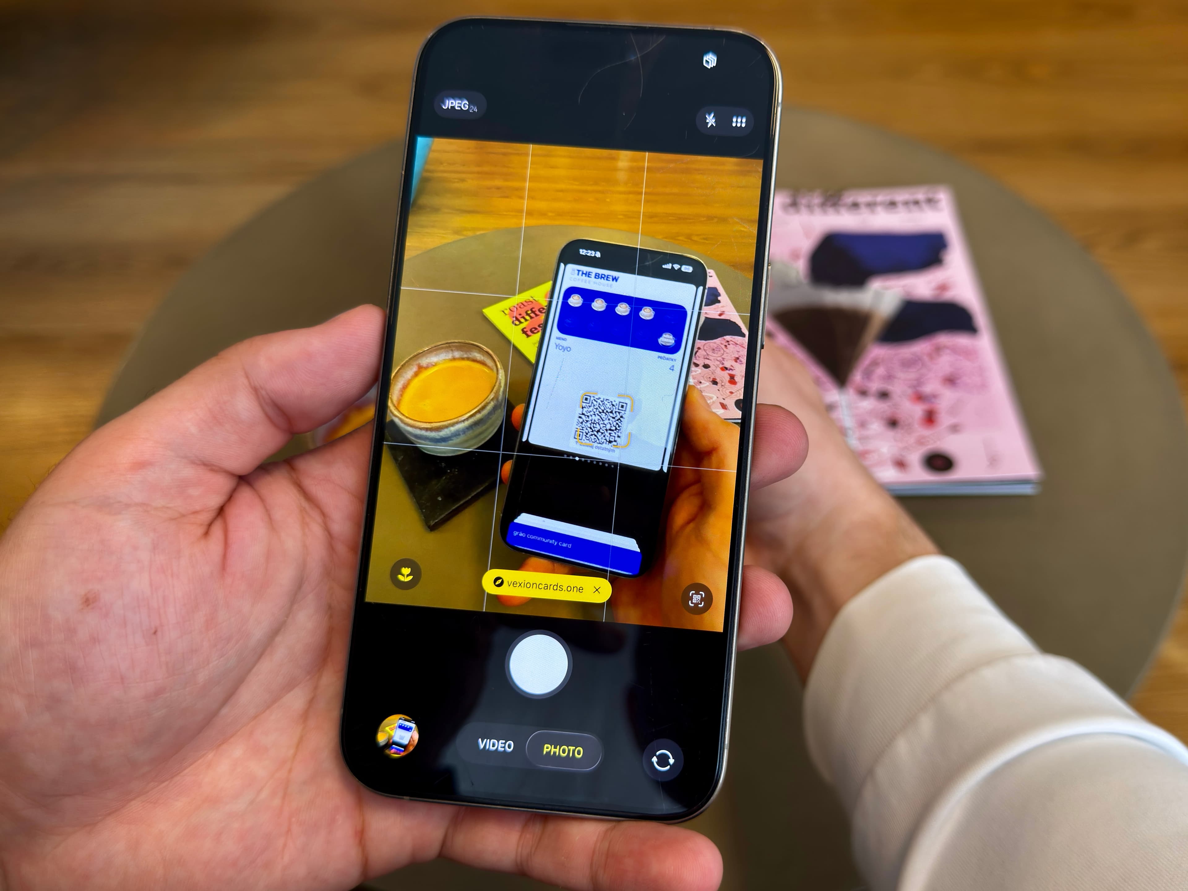 Ruka drží smartfón s otvorenou aplikáciou fotoaparátu, ktorá skenuje QR kód na digitálnej vernostnej karte do kaviarne zobrazený na inom telefóne na stole vedľa espressa a časopisov.
