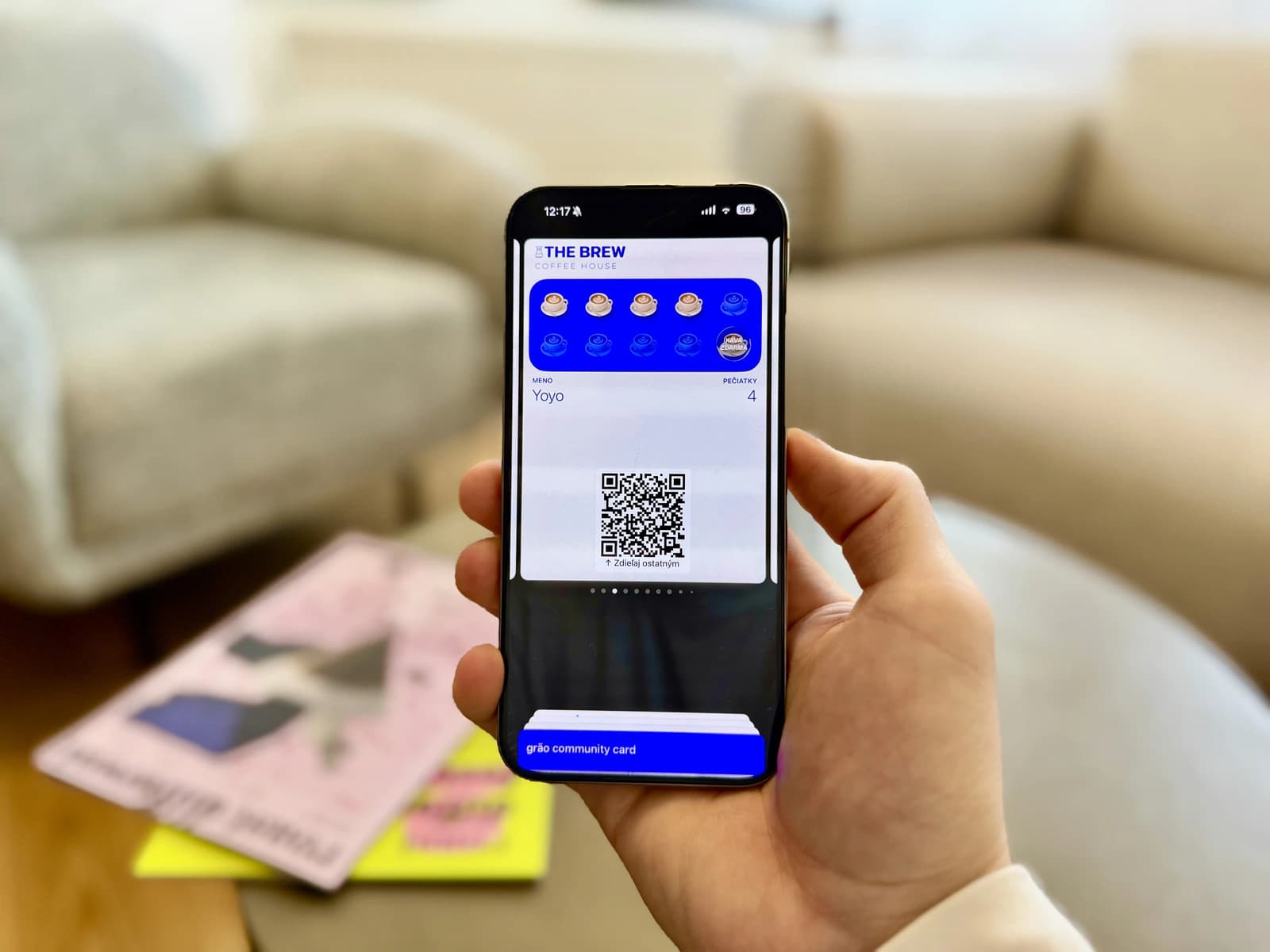 Ruka drží smartfón zobrazujúci digitálnu vernostnú kartu kaviarne s ikonami pečiatok a QR kódom, pričom v pozadí je rozmazaný stôl s časopismi.